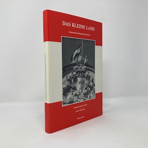 May include: A red and white book titled "Das Kleine Land" with the subtitle "Ein  buch  für  kleine  und  große  menschen". The cover features a black and white photo of a sculpture with a flag.