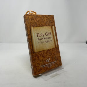 Peut inclure: Un livre à couverture rigide intitulé "Holy Gita Ready Reference" avec une couverture marron décorative. Le livre présente un design de parchemin avec du texte et un signet en ruban. Le livre est présenté sur un présentoir transparent.