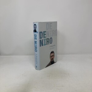 May include: A biography titled "De Niro" by John Baxter. The book has a light blue cover with the title in large, stylized letters. Images of the subject are featured on the cover. The book is standing upright.
