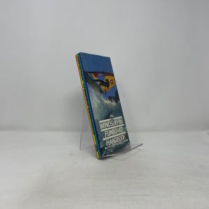 May include: A paperback book titled "The Windsurfing Funboard Handbook" with a blue cover featuring a windsurfer. The book's spine has a rainbow colour scheme. The book is propped up on a clear plastic stand.