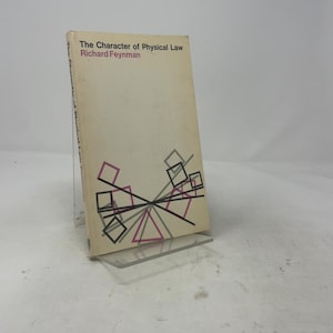 May include: A softcover book titled The Character of Physical Law by Richard Feynman. The off-white cover features black and magenta geometric shapes, including squares and triangles, arranged in a central pattern near the bottom edge.