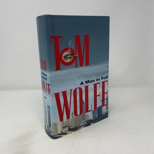 May include: A hardcover book titled A Man in Full by Tom Wolfe. The dust jacket features a blue sky background, a cityscape at the bottom, and large red text. A circular cutout in the letter O reveals a single human eye looking outward.
