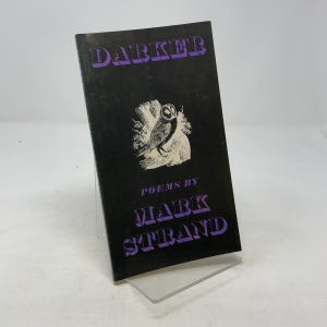 May include: A vintage book titled "Darker" by Mark Strand. The cover is black with purple text and an illustration of an owl. The book is displayed on a clear stand.