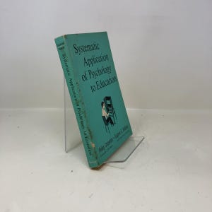 以下が含まれることがあります： 「Systematic Application of Psychology to Education」というタイトルのヴィンテージ本。ターコイズブルーの表紙で、コンピューターの前にいる人のグラフィックが特徴です。本は透明なスタンドに立てかけられています。