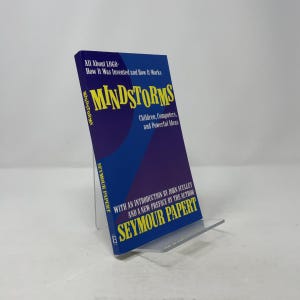 May include: A hardcover book titled "Mindstorms" by Seymour Papert, displayed on a clear acrylic stand. The book has a blue and purple cover with the title in large yellow letters. The text reads "All About LOGO - How It Was Invented and How It Works".