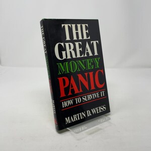 May include: A hardcover book titled "THE GREAT MONEY PANIC" with the subtitle "HOW TO SURVIVE IT" by Martin D. Weiss. The book has a black cover with white, green, and red text. The book is displayed on a clear plastic stand.