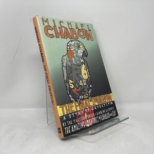 May include: A hardcover book titled "The Final Solution" by Michael Chabon. The cover features a colorful illustration of a figure composed of numbers and shapes. The book is displayed on a clear stand.