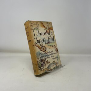 May include: A vintage book titled "I Traveled a Lonely Land" with illustrations of Australian animals and landmarks. The cover is tan with black and green text and illustrations. The book is propped up on a clear stand.