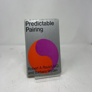 May include: A book titled "Predictable Pairing" with a silver cover. The cover features a yin-yang design in orange and purple. The authors' names, Robert A. Ravich, M.D. and Barbara Wyden, are printed below the design.