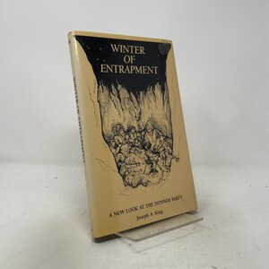 Könnte beinhalten: Ein gebundenes Buch mit dem Titel Winter of Entrapment: A New Look at the Donner Party von Joseph A. King. Der beigefarbene Schutzumschlag zeigt eine schwarze Tuschezeichnung einer Gruppe eng beieinander sitzender Menschen.