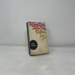 May include: A vintage paperback book by Tom Wolfe, titled "The Kandy-Kolored Tangerine-Flake Streamline Baby". The cover has colourful text in pink, orange, and yellow on a cream background. The book is propped up on a clear stand.