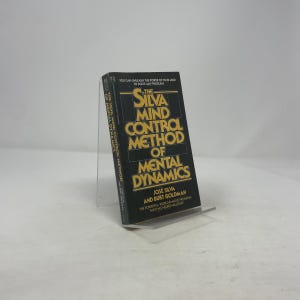May include: A hardcover book titled "The Silva Mind Control Method of Mental Dynamics" is displayed on a clear acrylic stand. The book has a black cover with gold lettering and the author's names, José Silva and Burt Goldman.