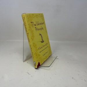 Könnte beinhalten: Ein Buch mit gelbem Einband mit dem Titel "The Velveteen Principles" und einer Zeichnung eines Kaninchens. Das Buch wird auf einem transparenten Acrylständer präsentiert. Buchrücken und Einband weisen Text auf.