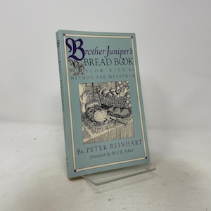 Peut inclure: Un livre relié bleu clair intitulé Brother Juniper's Bread Book: Slow Rise as Method and Metaphor par Br. Peter Reinhart, avec une préface de M.F.K. Fisher. La couverture présente un dessin au trait noir et blanc de pain et d'un panier.
