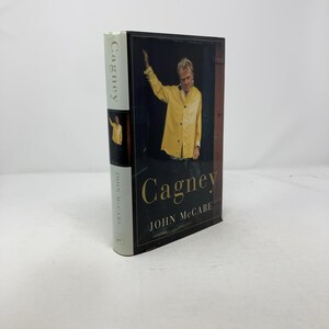 Könnte beinhalten: Ein gebundenes Buch mit dem Titel "Cagney" von John McCabe. Das Cover zeigt einen Mann in einer gelben Jacke. Der Buchrücken zeigt den Titel und den Namen des Autors vertikal. Schlichter Hintergrund.