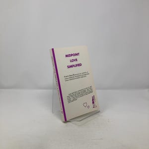 Puede incluir: Un libro de bolsillo titulado "MIDPOINT LOVE SIMPLIFIED" se muestra en un soporte acrílico transparente. El libro tiene una cubierta blanca con texto púrpura y un lomo púrpura. La portada también presenta una pequeña ilustración de dos personas y corazones.