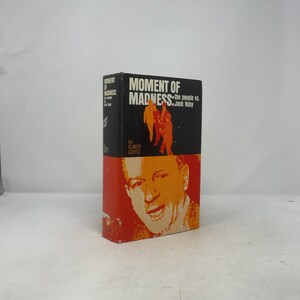 Puede incluir: Un libro titulado "Moment of Madness: The People vs. Jack Ruby" de Elmer Gertz. La portada está dividida en secciones negra y naranja, con figuras y texto naranja en la sección negra y un retrato en la sección naranja.