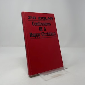 Puede incluir: Un libro de tapa dura rojo titulado "Confessions Of A Happy Christian" de Zig Ziglar. El título y el nombre del autor están en texto negro. El libro está apoyado en un soporte de plástico transparente sobre un fondo blanco.