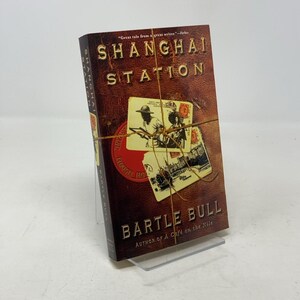 Op de afbeelding: Een hardcover boek met de titel "Shanghai Station" van Bartle Bull. De bruine kaft heeft de titel in gouden letters. Het boek is vastgebonden met touw en heeft een vintage ansichtkaartontwerp.