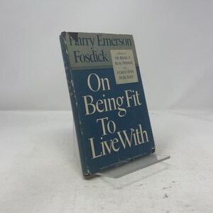 May include: A blue hardcover book by Harry Emerson Fosdick, titled "On Being Fit To Live With", displayed on a clear stand. The book's cover features white lettering and mentions "On Being A Real Person" and "A Great Time To Be Alive".