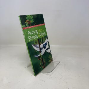 May include: A hardcover book titled "Pruning Trees, Shrubs & Vines" is displayed on a clear acrylic stand. The book cover is green with the title in white and the Brooklyn Botanic Garden logo. The cover features a close-up of pruning shears.