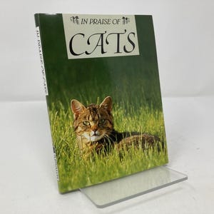 May include: A book titled "IN PRAISE OF CATS" featuring a tabby cat lying in green grass. The book is propped up on a clear acrylic stand. The book's cover has a green background with the title in large white letters.