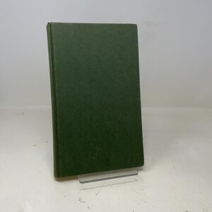 May include: A dark green hardcover book, standing on a clear plastic stand. The book's cover has a textured finish, giving it a classic appearance. The simple design suggests a vintage or classic literary work.