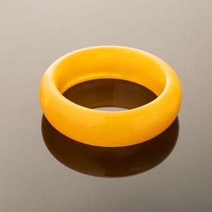 May include: A solid, smooth, circular bangle bracelet in a vibrant, translucent golden-yellow hue. The piece features a thick, rounded profile with a polished, glossy finish that catches the light, creating a clear reflection on the surface below.