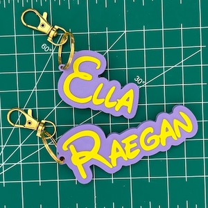 May include: Two personalized keychains with names "Ella" and "Raegan". The names are in yellow, outlined in purple, and attached to gold-colored clasps. The keychains are on a green cutting mat.