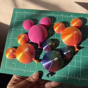 May include: A collection of colourful, balloon-shaped objects in shades of pink, orange, and purple, arranged on a green cutting mat. The balloons have a gradient effect, with a small stem at the bottom. The mat has inch measurements.