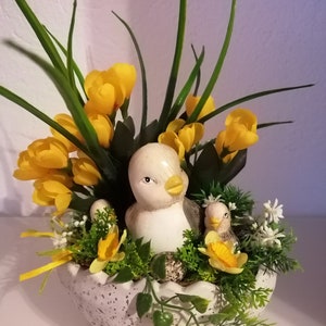 Könnte beinhalten: Eine weiße Keramikfigur eines Vogels und eines Küken, die in einem weißen, gefleckten, eiförmigen Topf eingebettet sind. Der Topf ist mit gelben Blumen, grünen Blättern und weißen Blüten gefüllt.