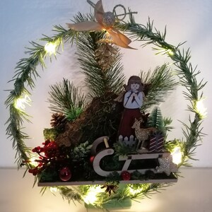 Könnte beinhalten: Ein festlicher Weihnachtskranz mit einem Holzengel, einem Hirsch und einem Schlitten. Der Kranz ist mit Tannennadeln, roten Beeren und kleinen Lichtern geschmückt. Oben befinden sich eine goldfarbene Schleife und ein Tannenzapfen. Die Basis ist eine Holzplattform.