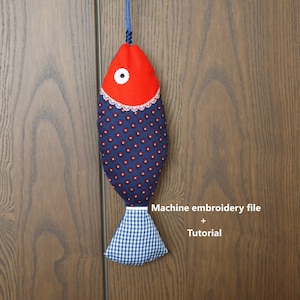 Peut inclure: Une pochette en tissu en forme de poisson rouge et bleu avec un œil blanc et une queue en vichy bleu et blanc. La pochette est suspendue à un cordon bleu et est étiquetée "Fichier de broderie machine + Tutoriel".