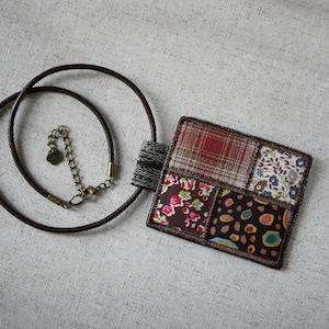 Op de afbeelding: Een ketting van bruin lederen koord met een vierkante hanger gemaakt van patchwork stof. De hanger heeft verschillende kleurrijke patronen, waaronder bloemen, ruitjes en geometrische ontwerpen.