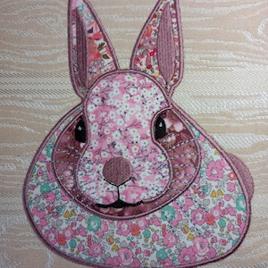 Peut inclure: Applique en tissu représentant un lapin avec un motif floral dans les tons de rose, blanc et bordeaux. Le lapin a de grandes oreilles, des yeux noirs et un petit nez. L'applique est bordée d'un fil rose plus foncé.