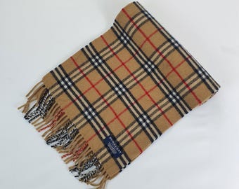 Op de afbeelding: Een beige sjaal met een klassiek ruitpatroon in rood, zwart en wit. De sjaal heeft een gefranjerde rand en een klein blauw label met de merknaam. De sjaal is opgevouwen en op een witte ondergrond geplaatst.