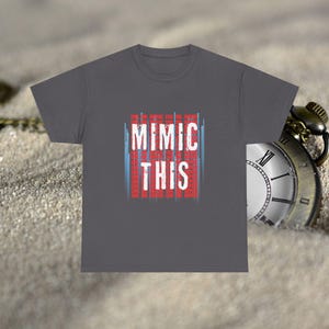 Pode incluir: T-shirt cinza escuro com o texto "MIMIC THIS" em branco, sobre um design retangular vermelho e azul. A camiseta é de estilo gola redonda.