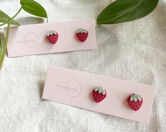 May include: Two pairs of red strawberry stud earrings on a pink card with the text "the playful clay" in a circle.