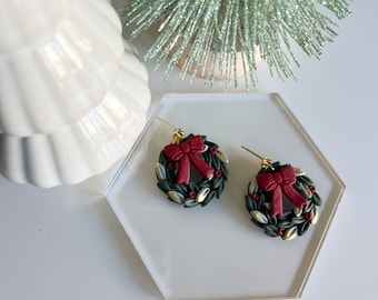 May include: A pair of green and gold Christmas wreath earrings with red bows. The earrings are on a white surface.