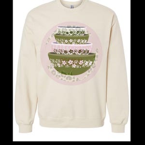 May include: A cream colored sweatshirt with a graphic of three stacked bowls with floral patterns in shades of green, pink, and white. The bowls are on a pink background with a leopard print.