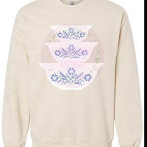 May include: A beige sweatshirt with a graphic design of three stacked bowls with floral patterns in shades of blue, pink, and white.