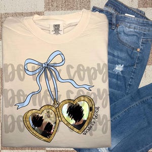 May include: Cream-colored Comfort Colors t-shirt with a gold heart-shaped locket pendant and a light blue bow. The locket is open, revealing a blurred image. Blue denim jeans are folded on the right side of the shirt.