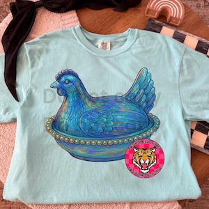 May include: A light blue Comfort Colors t-shirt featuring a detailed illustration of a blue hen sitting in a decorative bowl. The hen has intricate feather details and is surrounded by a beaded border. A round sticker with a tiger graphic is visible.