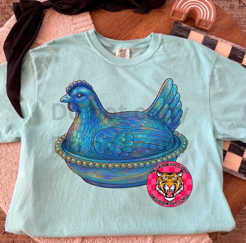 May include: A light blue Comfort Colors t-shirt featuring a detailed illustration of a blue hen sitting in a decorative bowl. The hen has intricate feather details and is surrounded by a beaded border. A round sticker with a tiger graphic is visible.