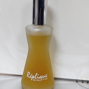 May include: A clear glass bottle of Replique spray mist. The bottle is shaped like an hourglass and has a silver cap. The label reads "Replique spray mist" in white lettering.