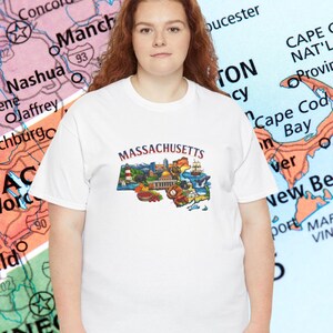May include: A white t-shirt featuring a colorful graphic of Massachusetts landmarks and the state name in maroon. The shirt is worn by a person standing in front of a map of New England.