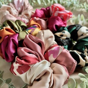 May include: A collection of colourful satin scrunchies in various patterns, including floral, polka dot, and solid colours. The scrunchies are arranged in a pile, with some overlapping.