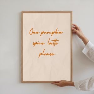 Może przedstawiać: Oprawiony wydruk z napisem "One pumpkin spice latte please" w pomarańczowym skrypcie. Wydruk ma kremowe tło i jasnobrązową drewnianą ramę. Obraz jest trzymany przez osobę noszącą białą koszulę.