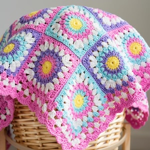 May include: A vibrant, hand-crocheted blanket with a floral design. The blanket displays squares in pink, purple, blue, yellow, and white. It is draped over a wicker basket, highlighting the detailed stitching and bright colours. The blanket is approximately 120cm x 120cm.