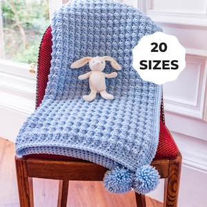 Puede incluir: Una manta de crochet azul claro con un patrón texturizado y dos pompones, sobre una silla de madera. Un pequeño conejito de peluche está sentado en la manta. La imagen incluye el texto "20 SIZES".